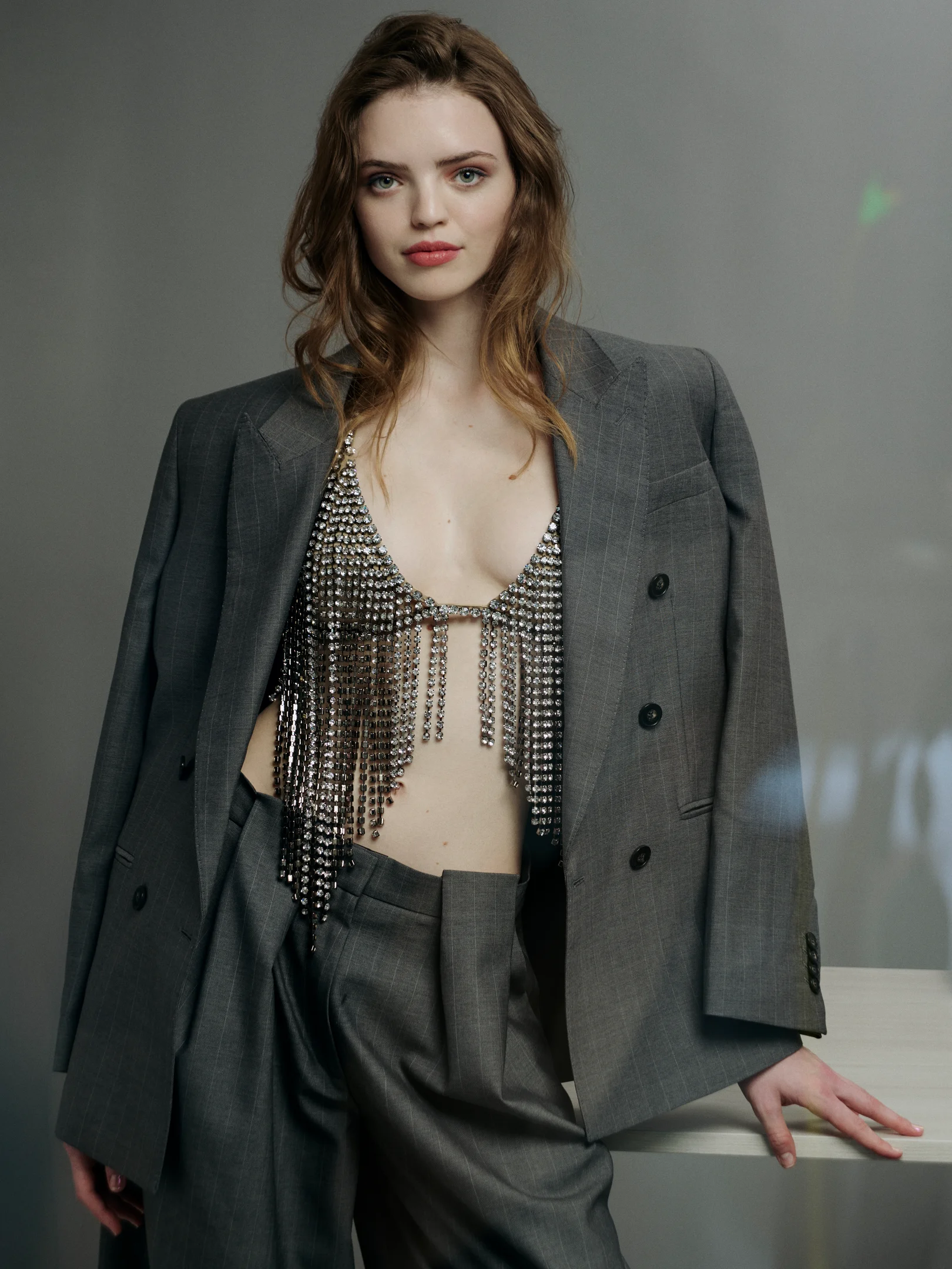 Frau in oversized grauem Anzug mit funkelndem, fransenbesetztem Top, frontal vor neutralem Hintergrund fotografiert
