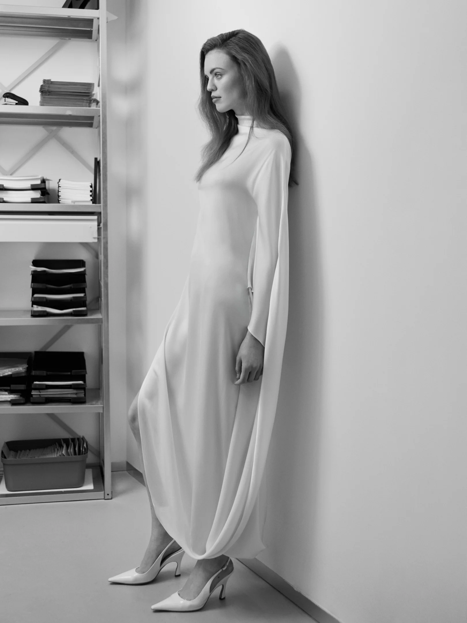 Schwarzweißaufnahme einer Frau in fließendem weißen Kleid, die an einer Wand neben einem Regal lehnt