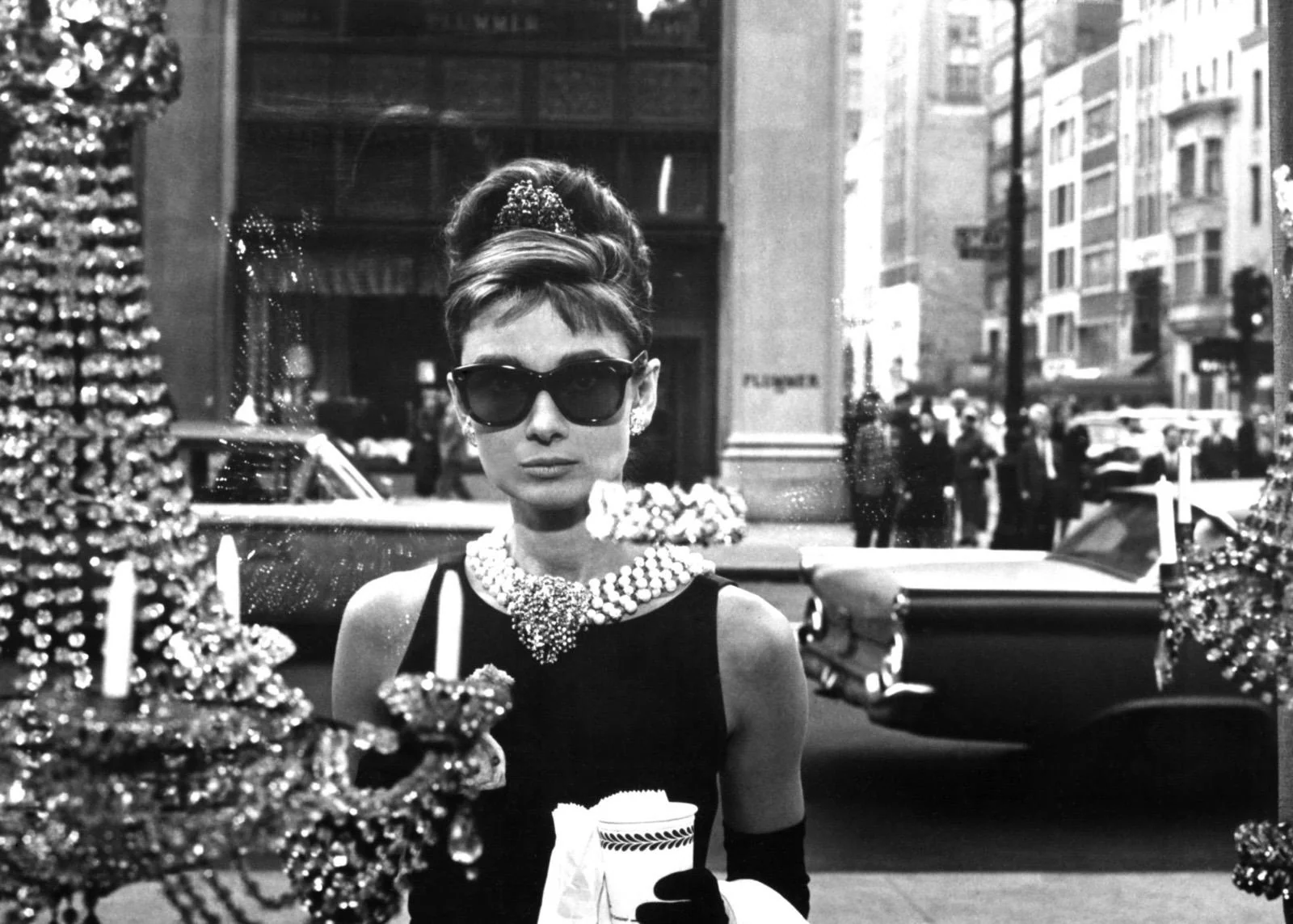 Schwarz-Weiß-Fotografie von Audrey Hepburn als Holly Golightly vor dem Tiffany-Schaufenster in New York mit Sonnenbrille und Kaffeebecher.