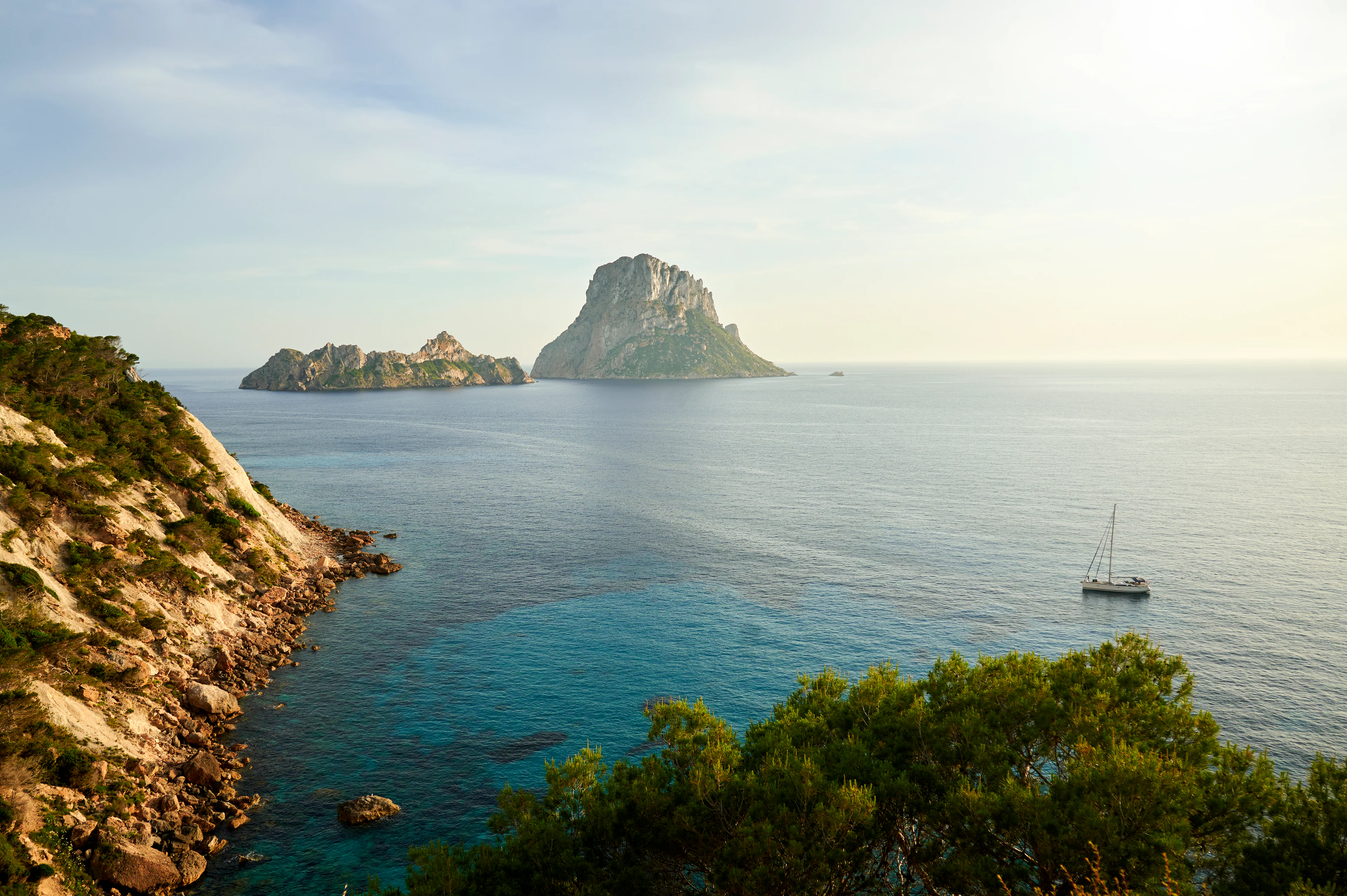 Blick auf die Felseninsel Es Vedrà vor der Küste Ibizas mit türkisblauem Mittelmeer und Segelboot – ein Highlight ausgewählter Explora Journeys Routen.