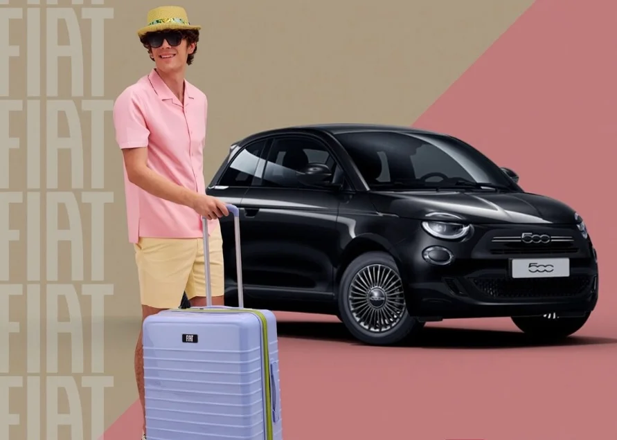 Model mit blauem Reisekoffer aus der Fiat x Ful Kollektion vor schwarzem Fiat 500.