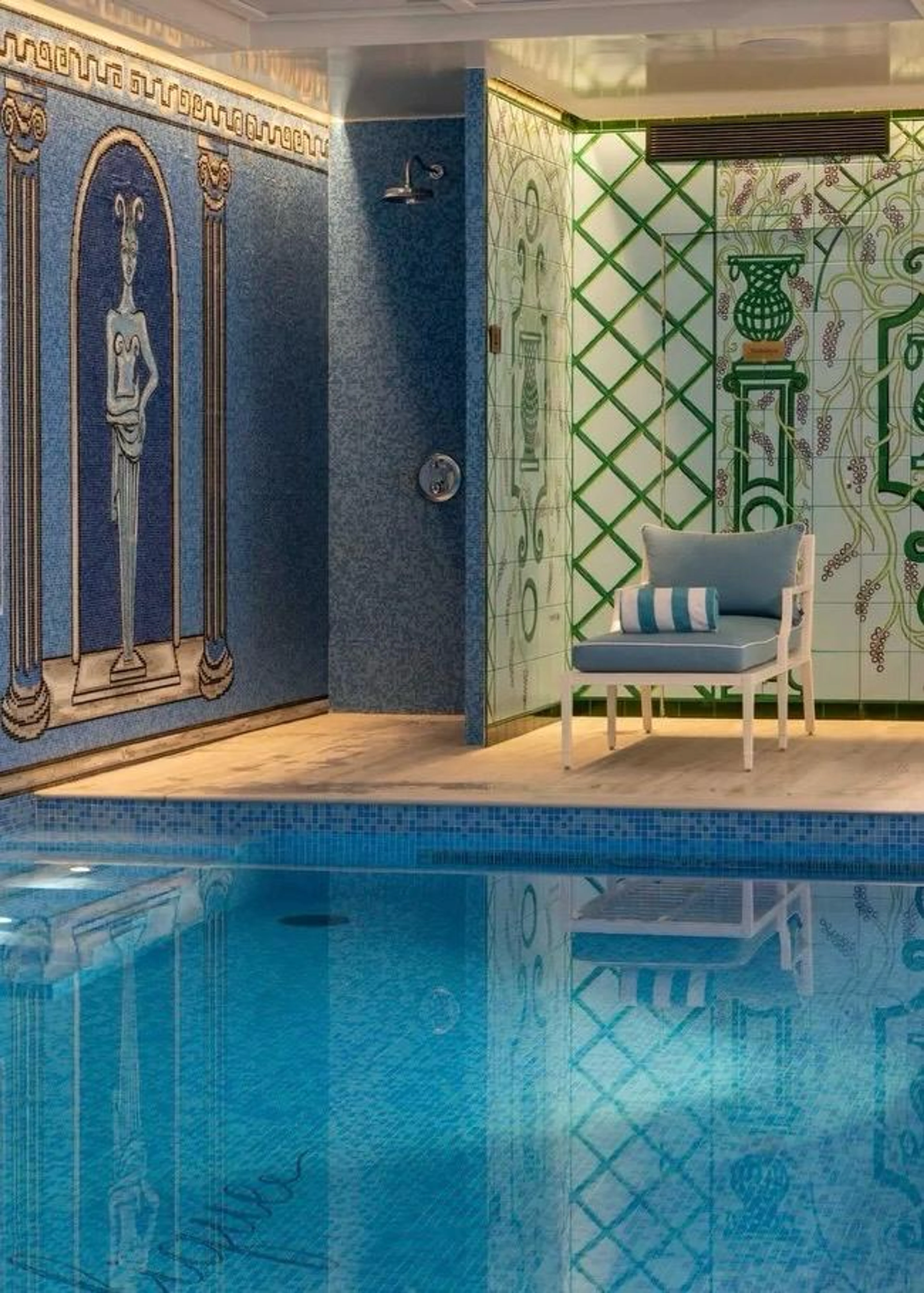 Der luxuriöse Hotel-Pool im Untergeschoss des Le Magnifique mit blauem Mosaik und antiken Figuren an der Wand sowie zwei hellblauen Liegestühlen vor grünem Gitterwerk-Dekor.