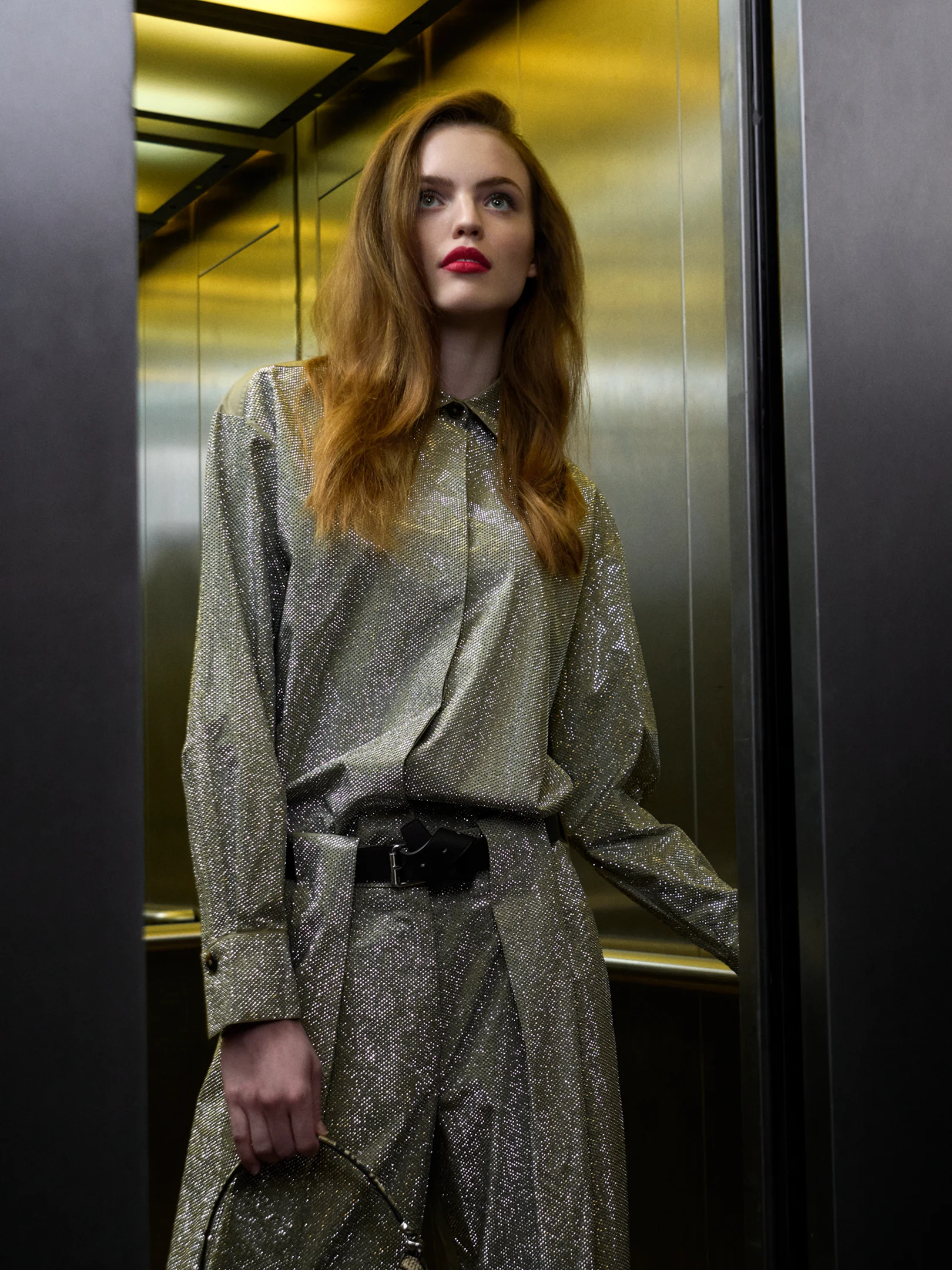 Frau in schimmerndem silbernen Outfit steht in einem Aufzug mit warmem Licht und blickt nach oben
