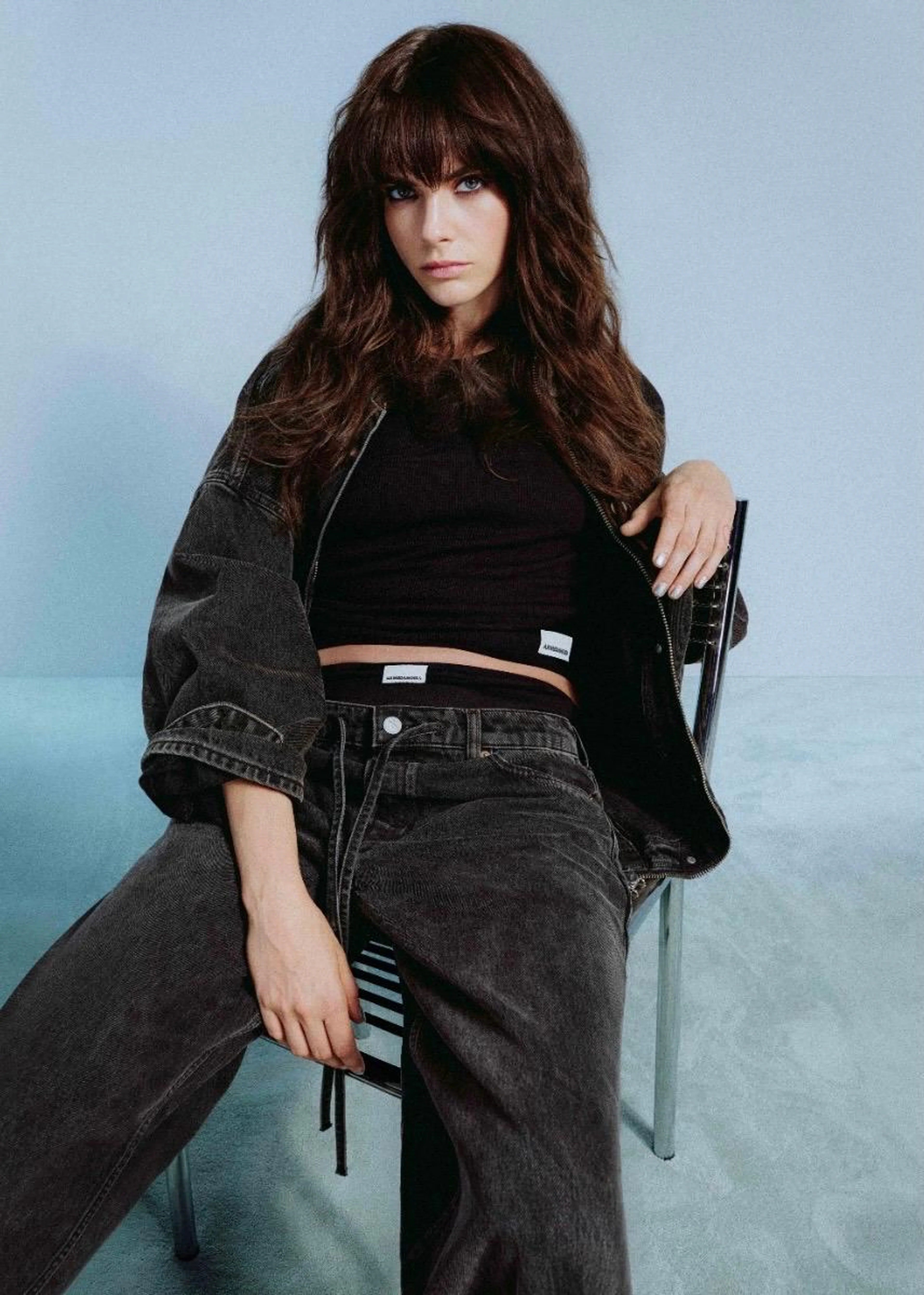 Model Cara Delevingne sitzt lässig auf einem Stuhl in einem schwarzen Denim-Outfit aus der Armedangels Detoxdenim-Kollektion.