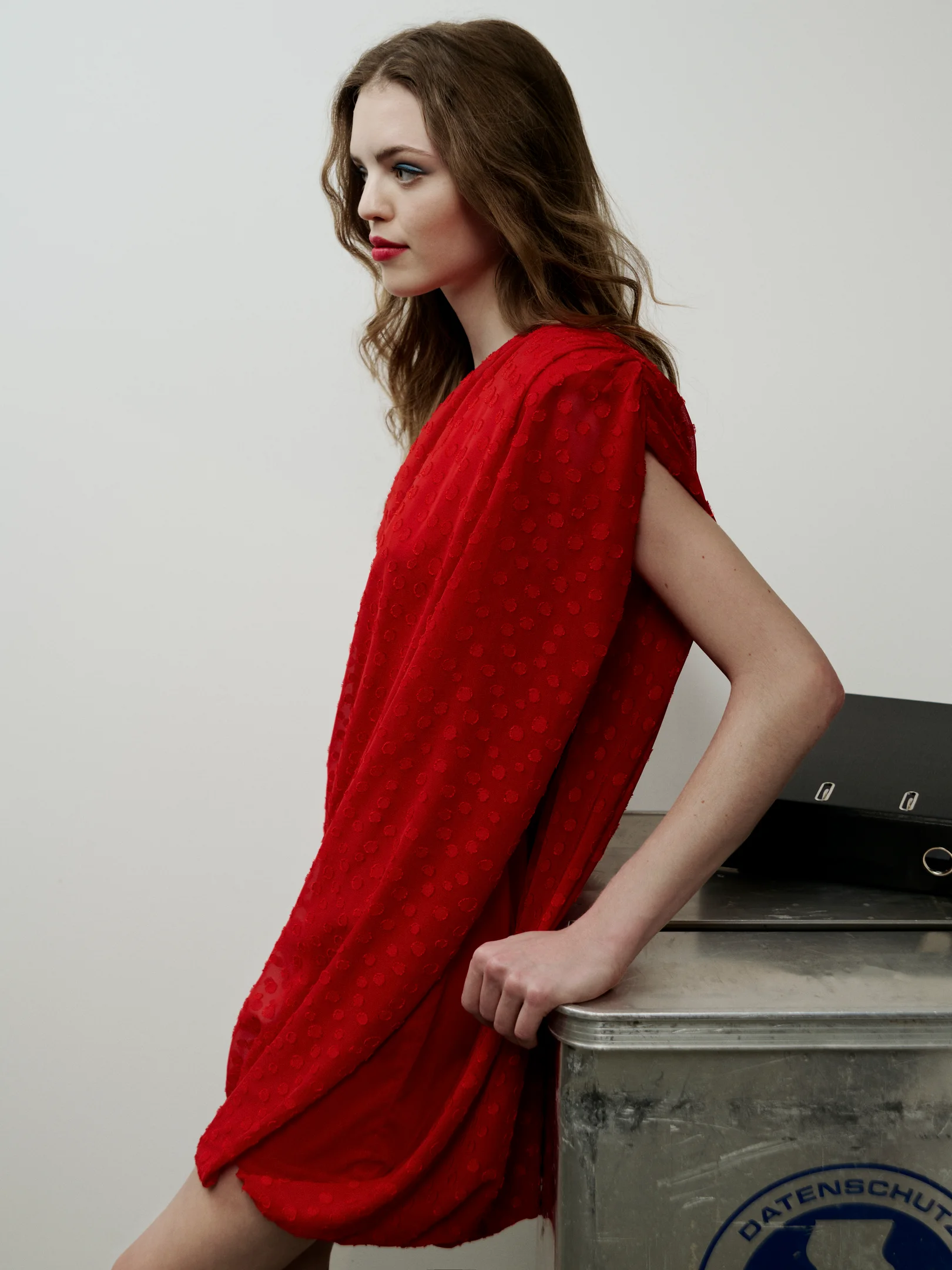 Frau im roten drapierten Kleid lehnt seitlich an einem Metalltisch in minimalistischem Raum