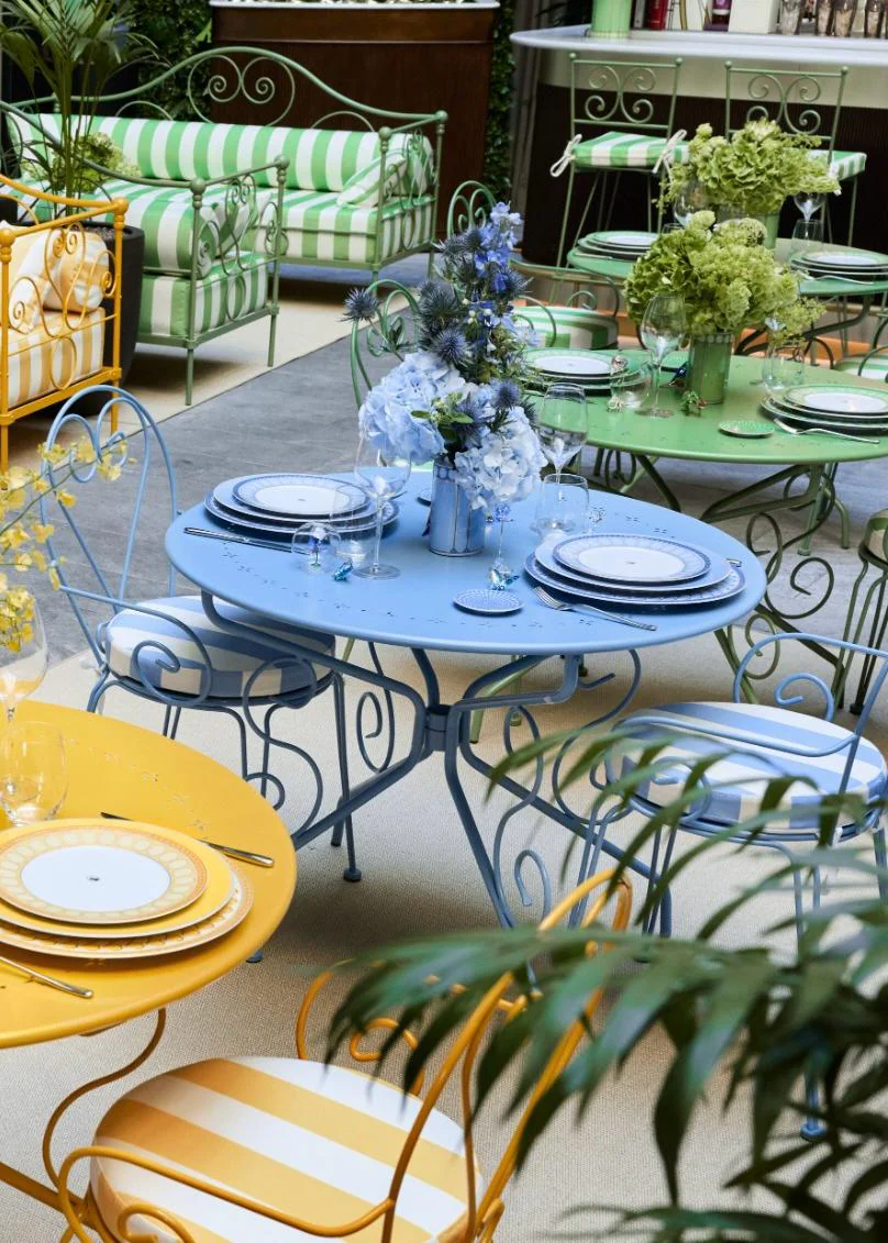 Der farbenfrohe Innenhof des Café Swarovski im Giardino Cordusio in Mailand Ein elegant gestalteter Innenhof mit verschnörkelten Metallmöbeln in Pastellfarben. Im Vordergrund stehen gelbe und blaue runde Tische mit eingedecktem Rosenthal x Swarovski Porzellan, im Hintergrund sind rosa, gelb und grün gestreifte Sofas zu sehen.