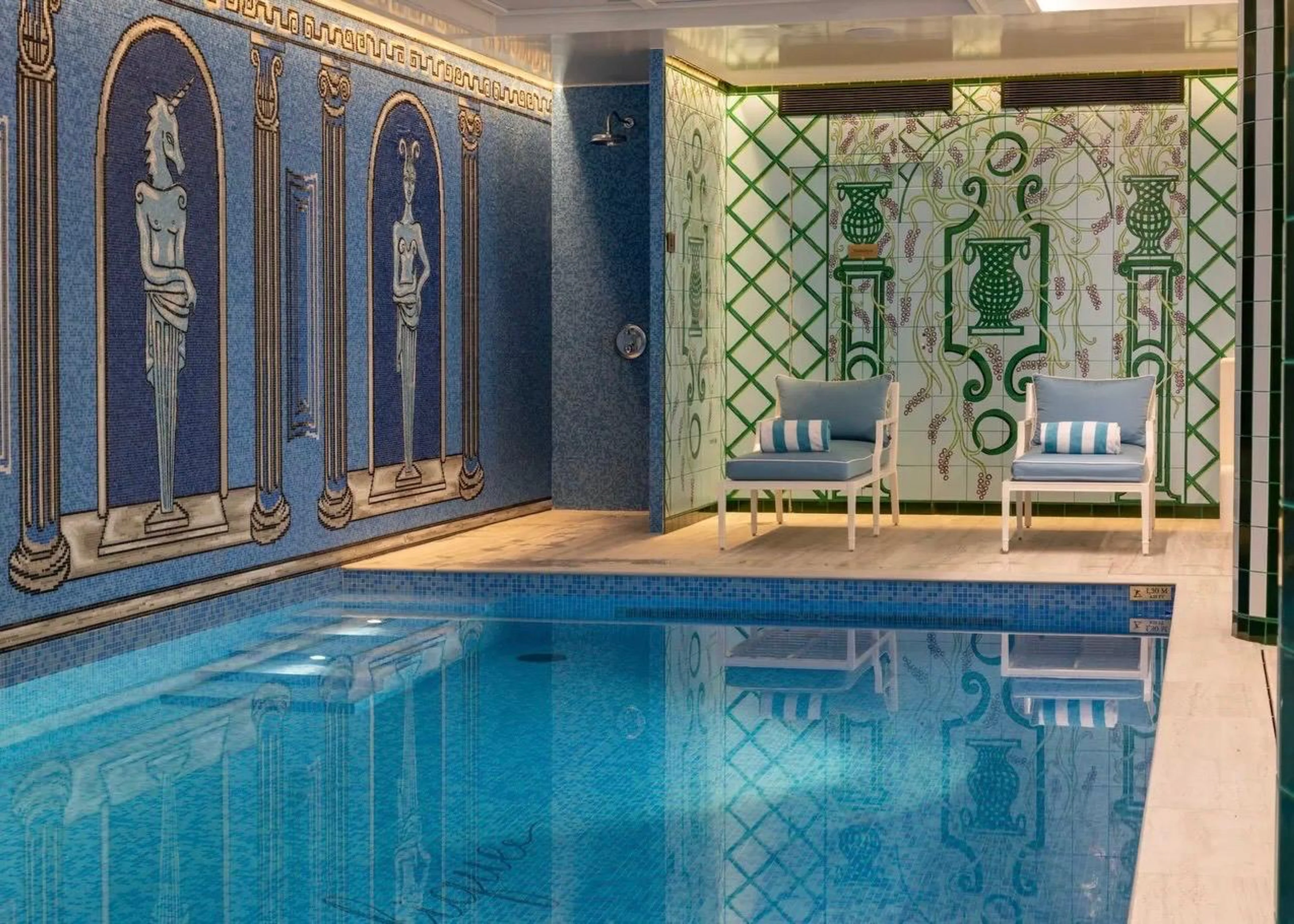 Der luxuriöse Hotel-Pool im Untergeschoss des Le Magnifique mit blauem Mosaik und antiken Figuren an der Wand sowie zwei hellblauen Liegestühlen vor grünem Gitterwerk-Dekor.