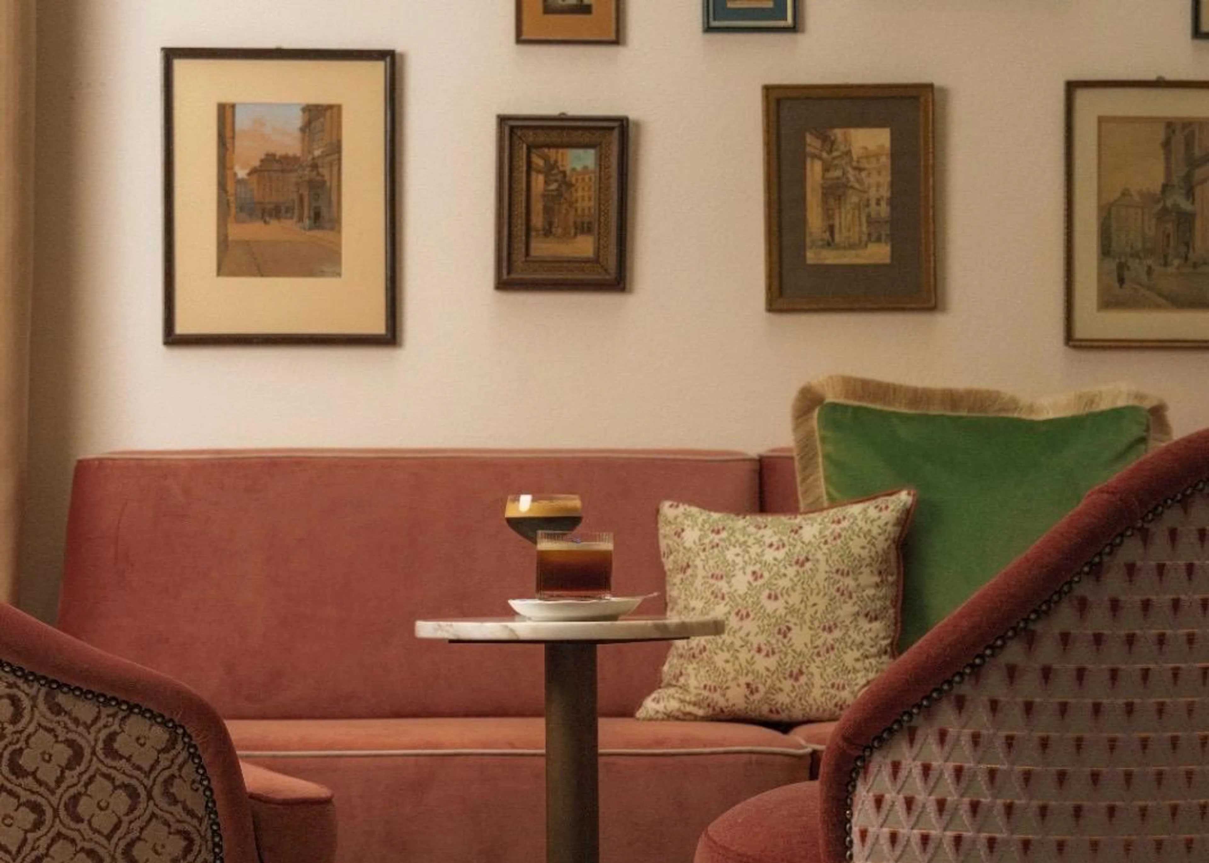 Eine gemütliche Kaffeeecke im Café Constanze mit einer rosa Samtcouch, Kissen, Bildern an der Wand und Kaffeespezialitäten auf einem Marmortisch.
