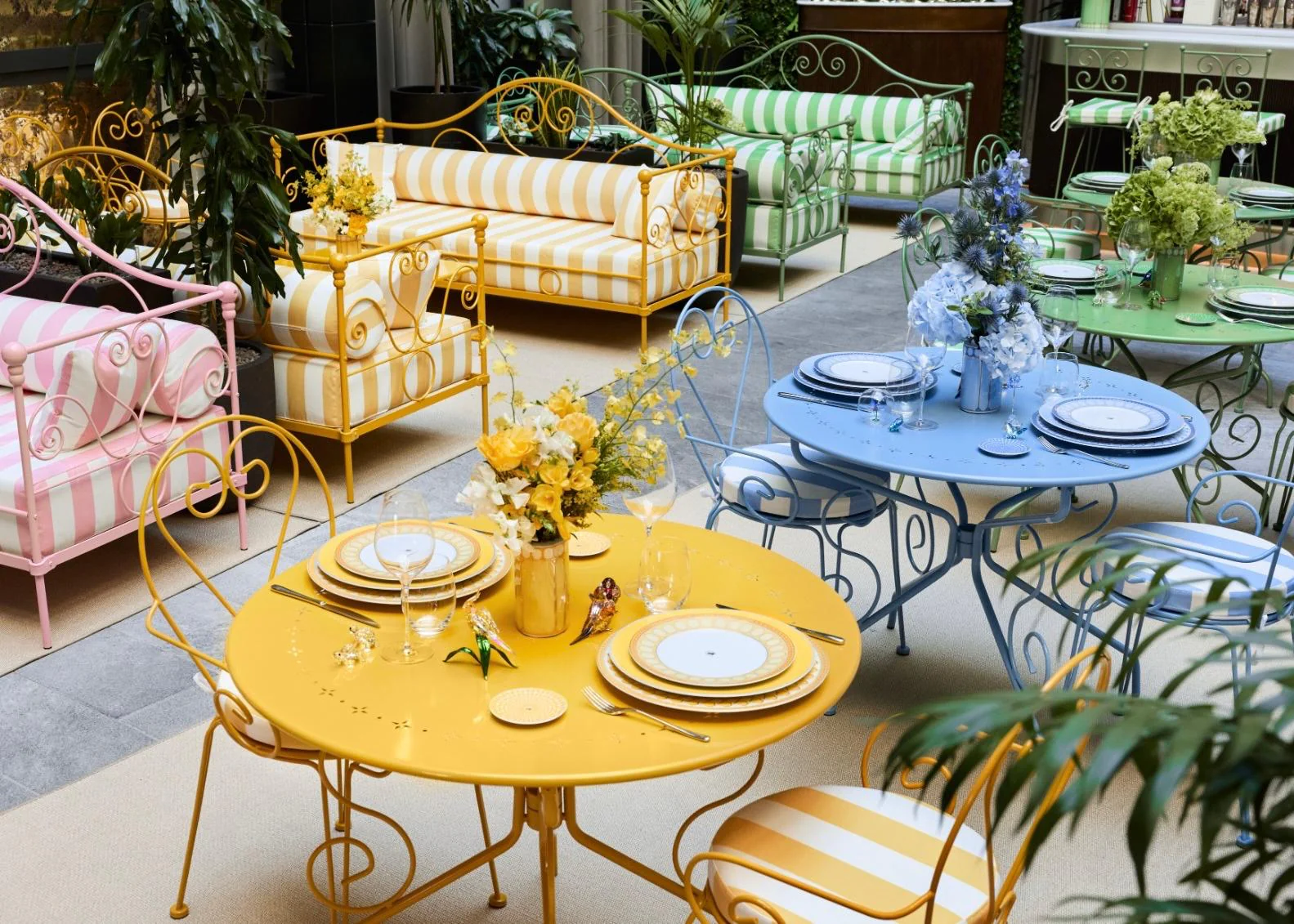 Ein elegant gestalteter Innenhof mit verschnörkelten Metallmöbeln in Pastellfarben. Im Vordergrund stehen gelbe und blaue runde Tische mit eingedecktem Rosenthal x Swarovski Porzellan, im Hintergrund sind rosa, gelb und grün gestreifte Sofas zu sehen.