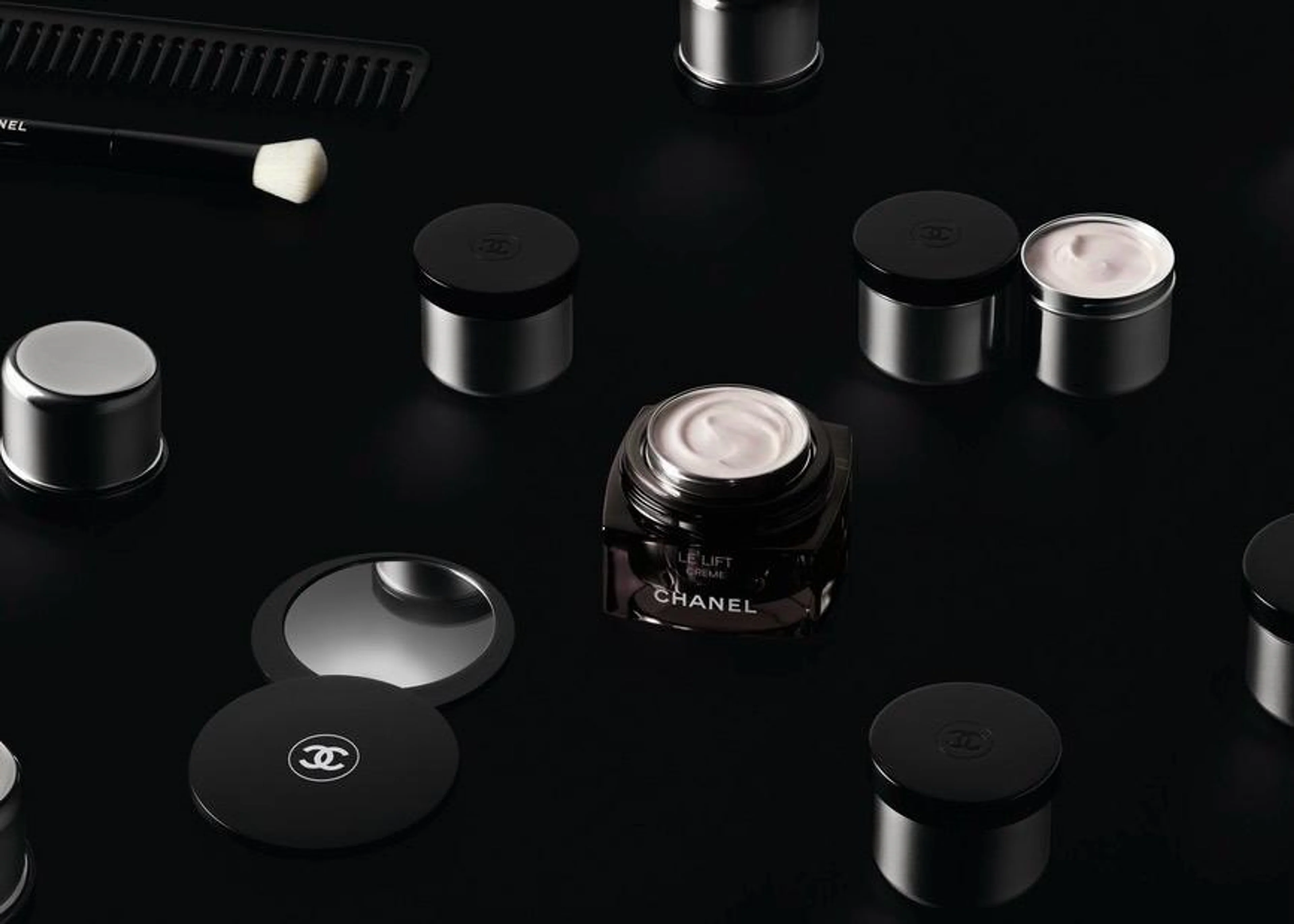 Geöffneter Tiegel der CHANEL LE LIFT Crème mit sichtbarer Textur inmitten von luxuriösen Kosmetikprodukten und Pinseln auf einer schwarzen, glänzenden Oberfläche.