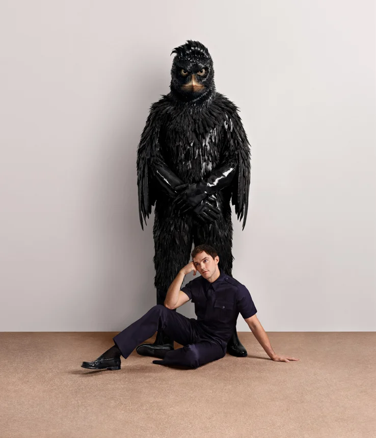 Männliches Model in dunklem Outfit sitzt vor großer schwarzer Vogel-Figur aus der Prada Kampagne