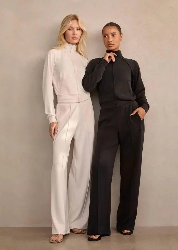 Georgina Rodríguez und ein blondes Model posieren nebeneinander vor einer grauen Wand. Sie tragen minimalistische Mimoa Trainingsanzüge mit Reißverschlussjacke und passender Sporthose in Schwarz und Cremeweiß.