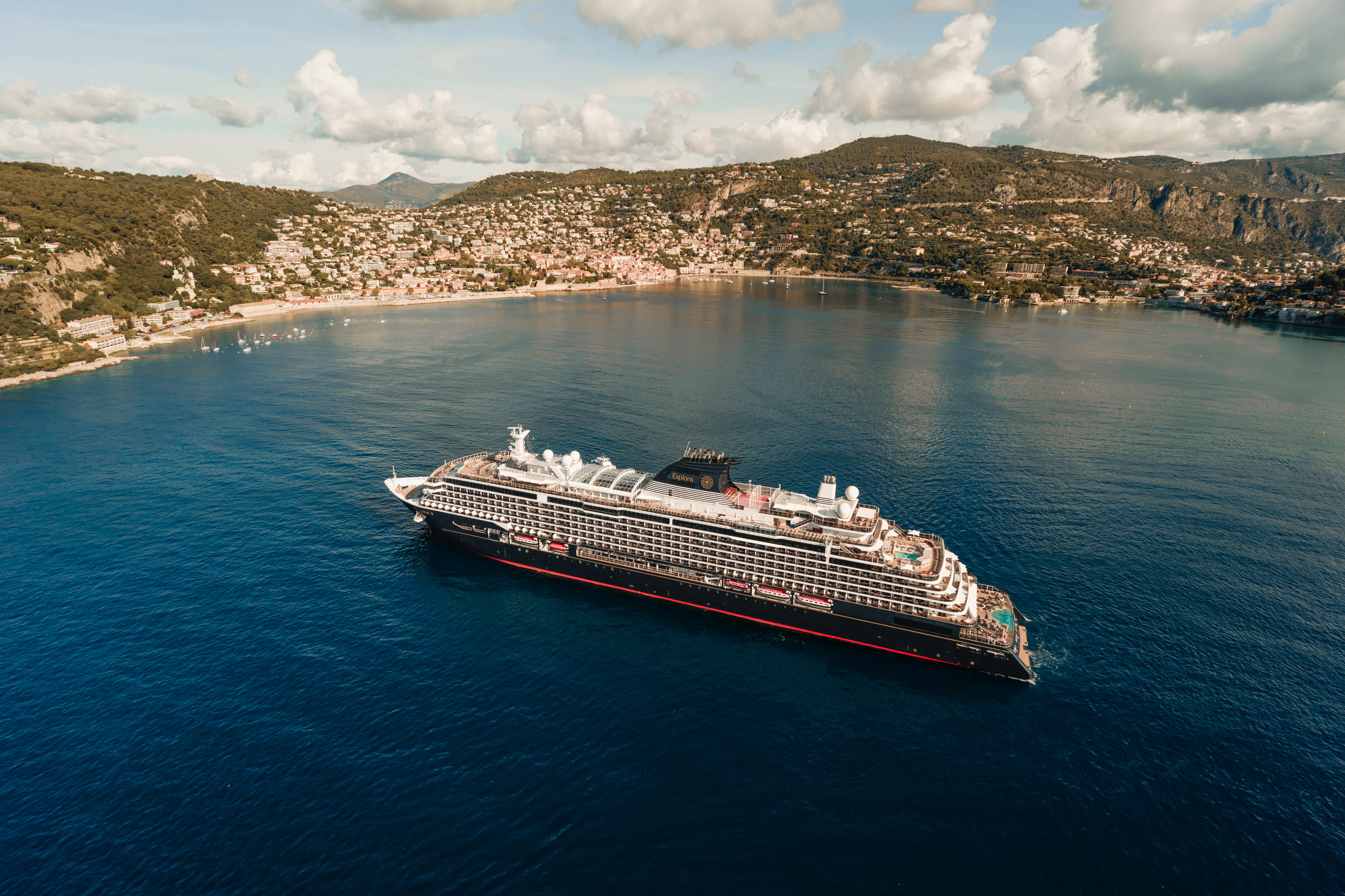 Die EXPLORA III von Explora Journeys auf ihrer Route durch das Mittelmeer vor einer mediterranen Küstenlandschaft.