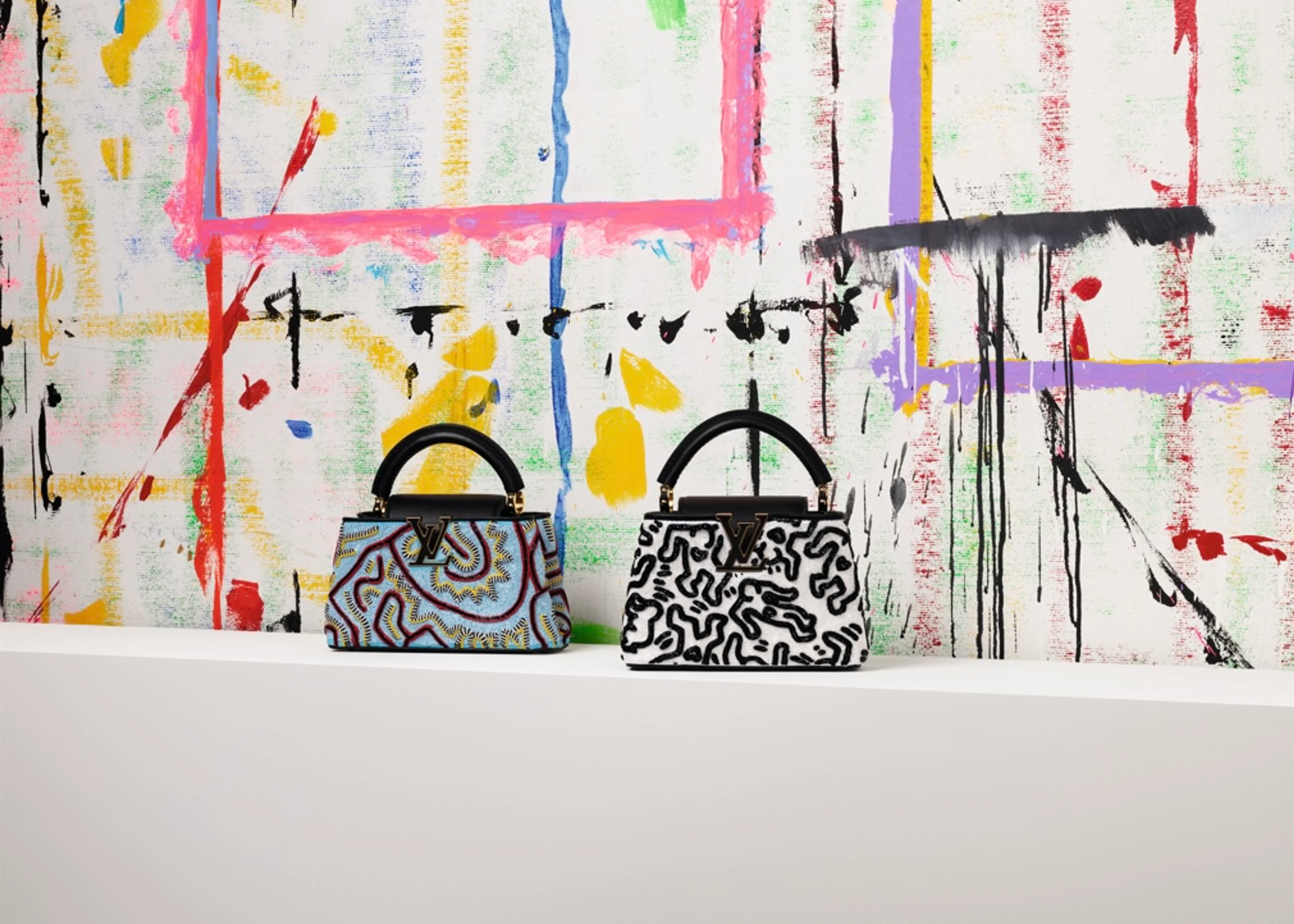 Zwei exklusive Louis Vuitton Capucines Handtaschen mit den ikonischen, abstrakten Mustern des Künstlers Keith Haring stehen auf einem weißen Tresen vor einer bunt bemalten Kunstleinwand.