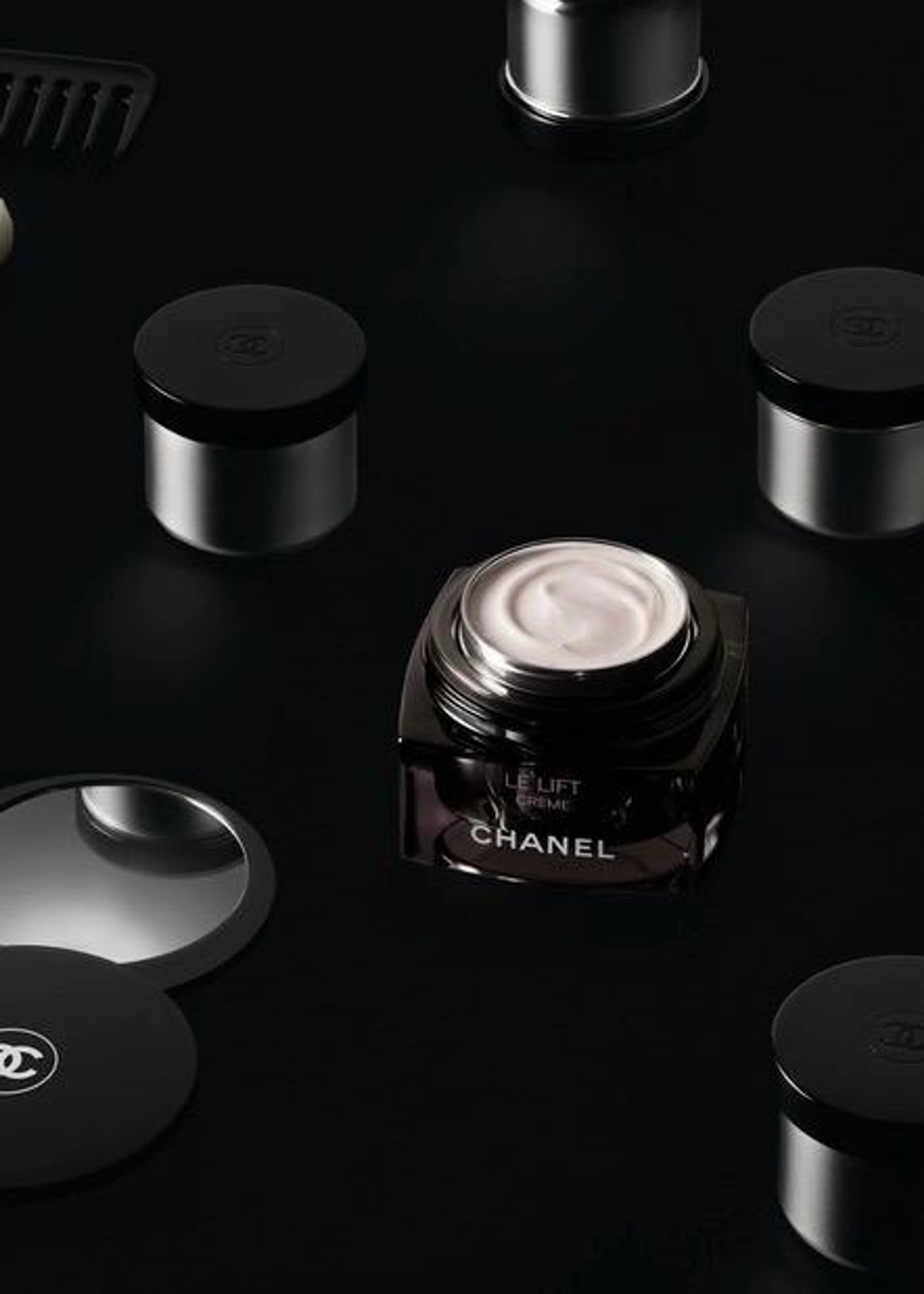Geöffneter Tiegel der CHANEL LE LIFT Crème mit sichtbarer Textur inmitten von luxuriösen Kosmetikprodukten und Pinseln auf einer schwarzen, glänzenden Oberfläche.