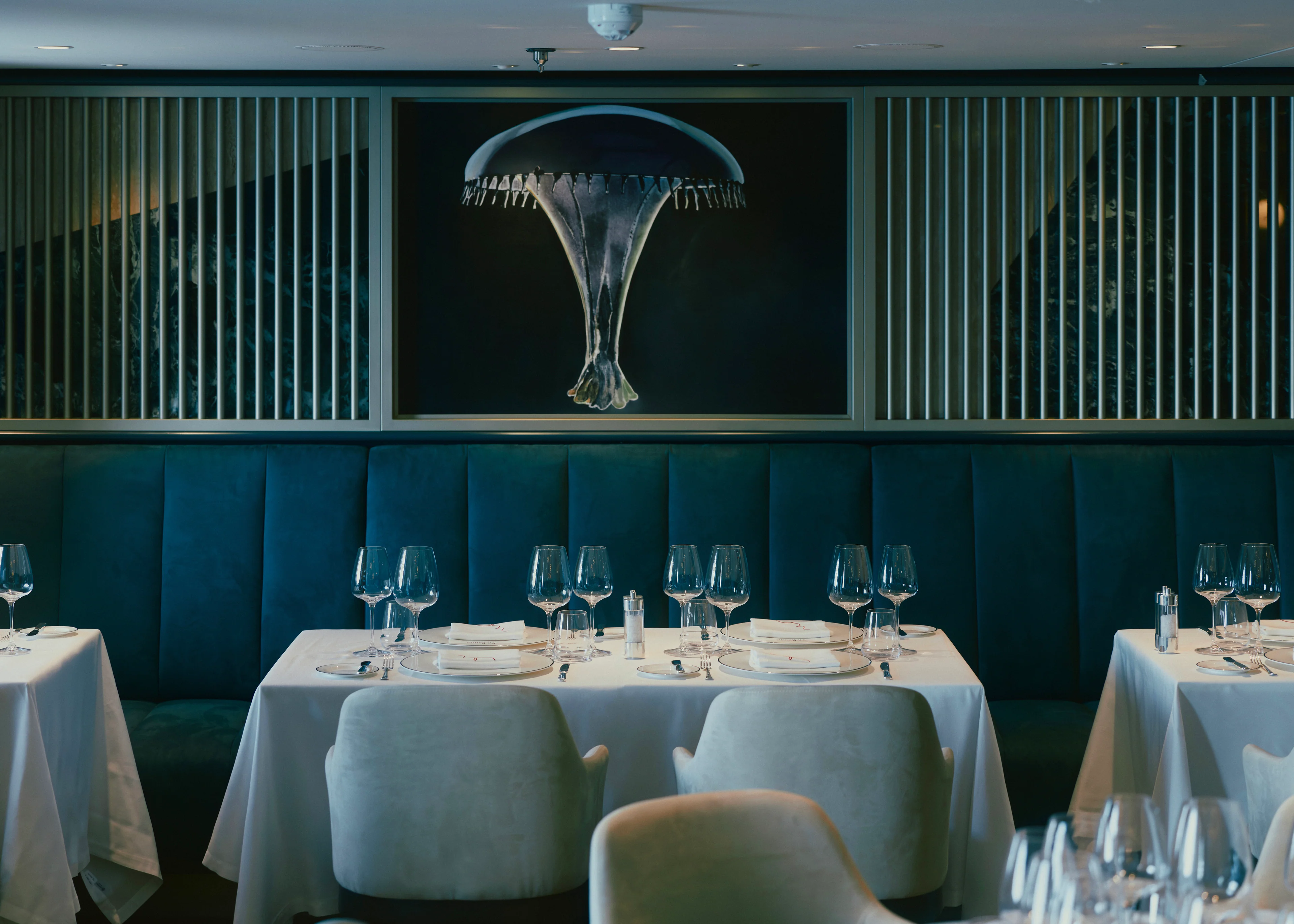Elegant eingedeckte Tische mit weißen Tischtüchern und Weingläsern im luxuriösen Restaurant Fil Rouge an Bord des Kreuzfahrtschiffs Explora III vor einer blauen Samt-Sitzbank und einem modernen Kunstwerk mit Quallen-Motiv.