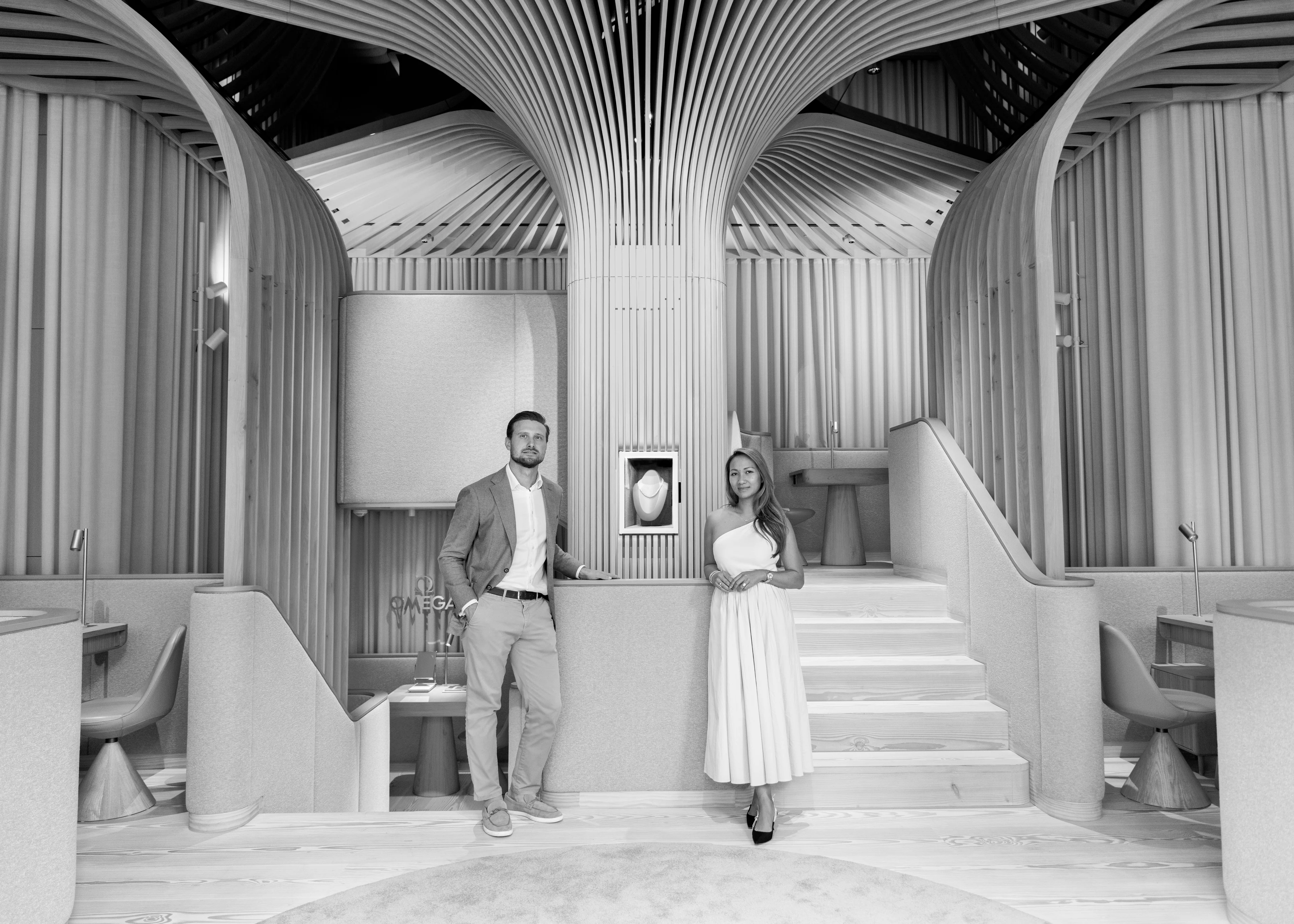 Schwarz-Weiß-Fotografie von Felix und Nattaya Köck, die elegant gekleidet in ihrer modern gestalteten Schmuckboutique mit geschwungenen Holzpaneelen neben einer beleuchteten Vitrine stehen.