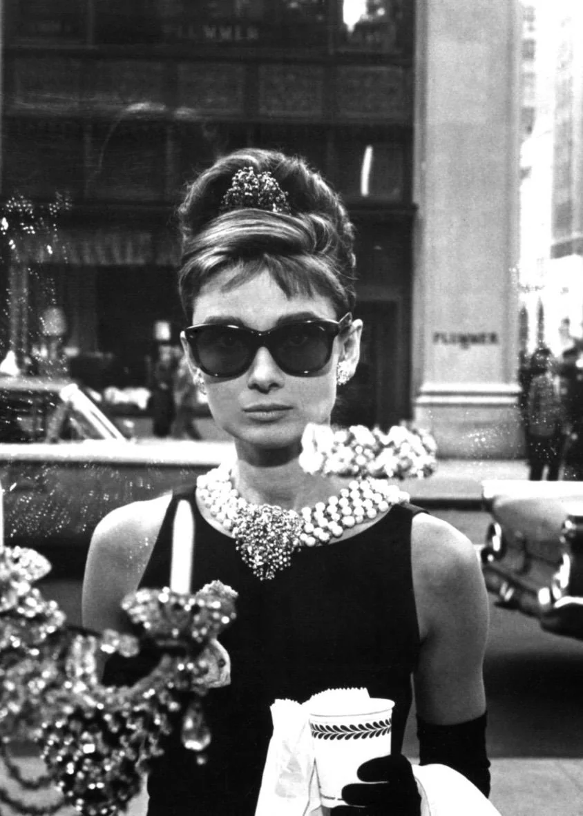 Schwarz-Weiß-Fotografie von Audrey Hepburn als Holly Golightly vor dem Tiffany-Schaufenster in New York mit Sonnenbrille und Kaffeebecher.