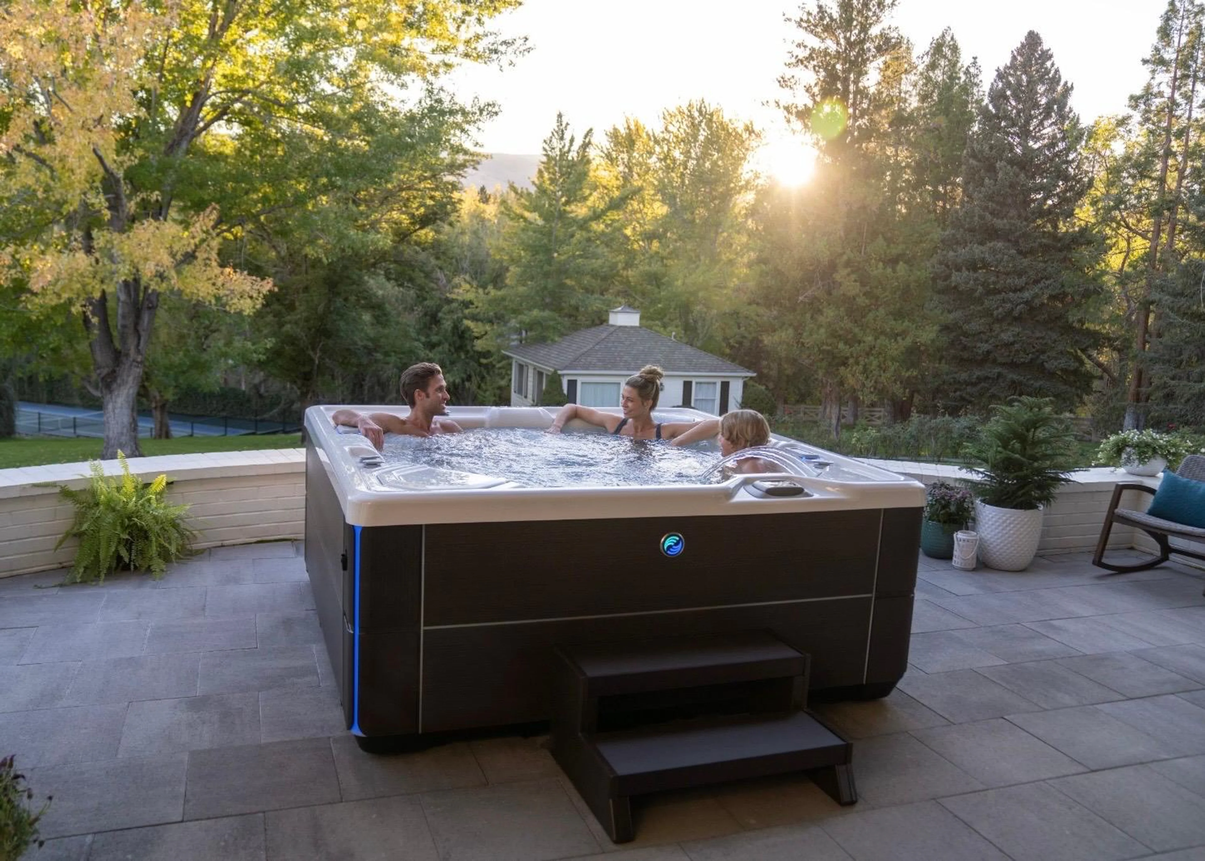 Drei Personen entspannen in einem modernen HotSpring Whirlpool auf einer gepflasterten Terrasse im Garten bei tiefstehender Abendsonne.