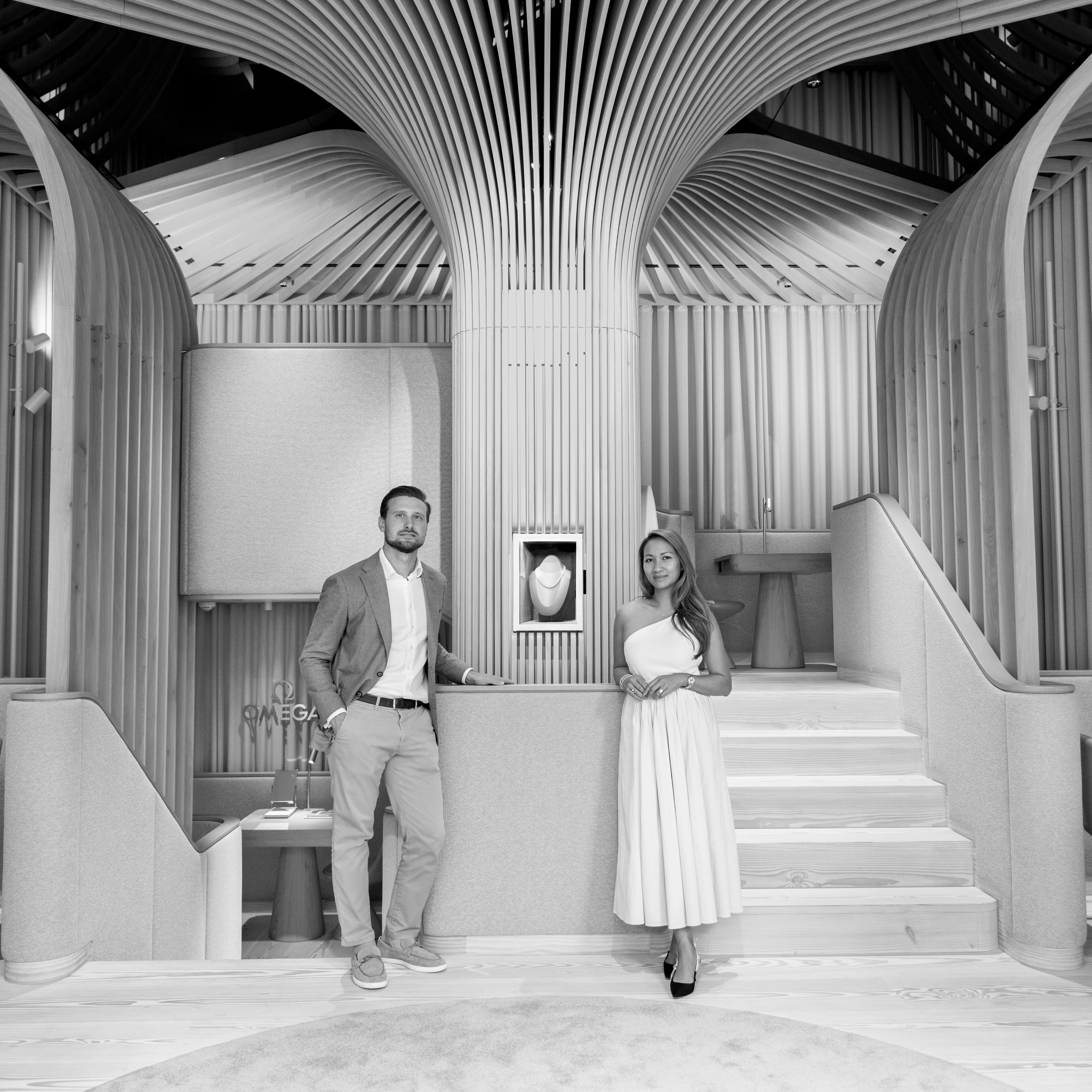 Schwarz-Weiß-Fotografie von Felix und Nattaya Köck, die elegant gekleidet in ihrer modern gestalteten Schmuckboutique mit geschwungenen Holzpaneelen neben einer beleuchteten Vitrine stehen.