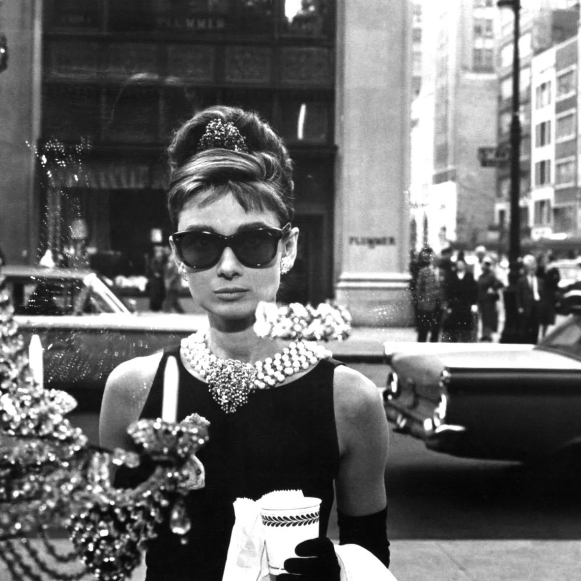 Audrey Hepburn in Frühstück bei Tiffany (1961) Schwarz-Weiß-Fotografie von Audrey Hepburn als Holly Golightly vor dem Tiffany-Schaufenster in New York mit Sonnenbrille und Kaffeebecher.