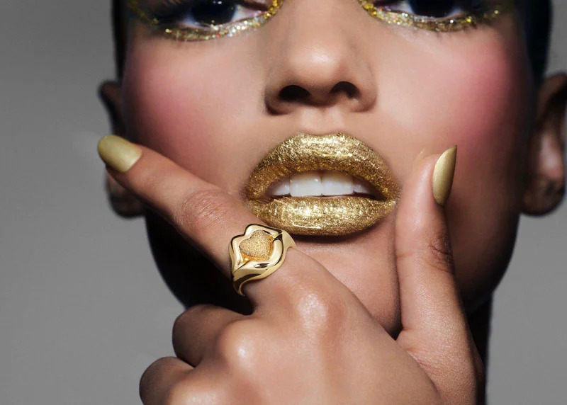 Nahaufnahme einer Frau mit glitzerndem, goldenem Lippenstift und Lidschatten, die einen auffälligen goldenen Siegelring in Form von Lippen mit einem Herz an der Hand trägt.