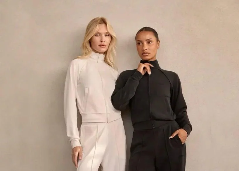 Georgina Rodríguez und ein blondes Model posieren nebeneinander vor einer grauen Wand. Sie tragen minimalistische Mimoa Trainingsanzüge mit Reißverschlussjacke und passender Sporthose in Schwarz und Cremeweiß.