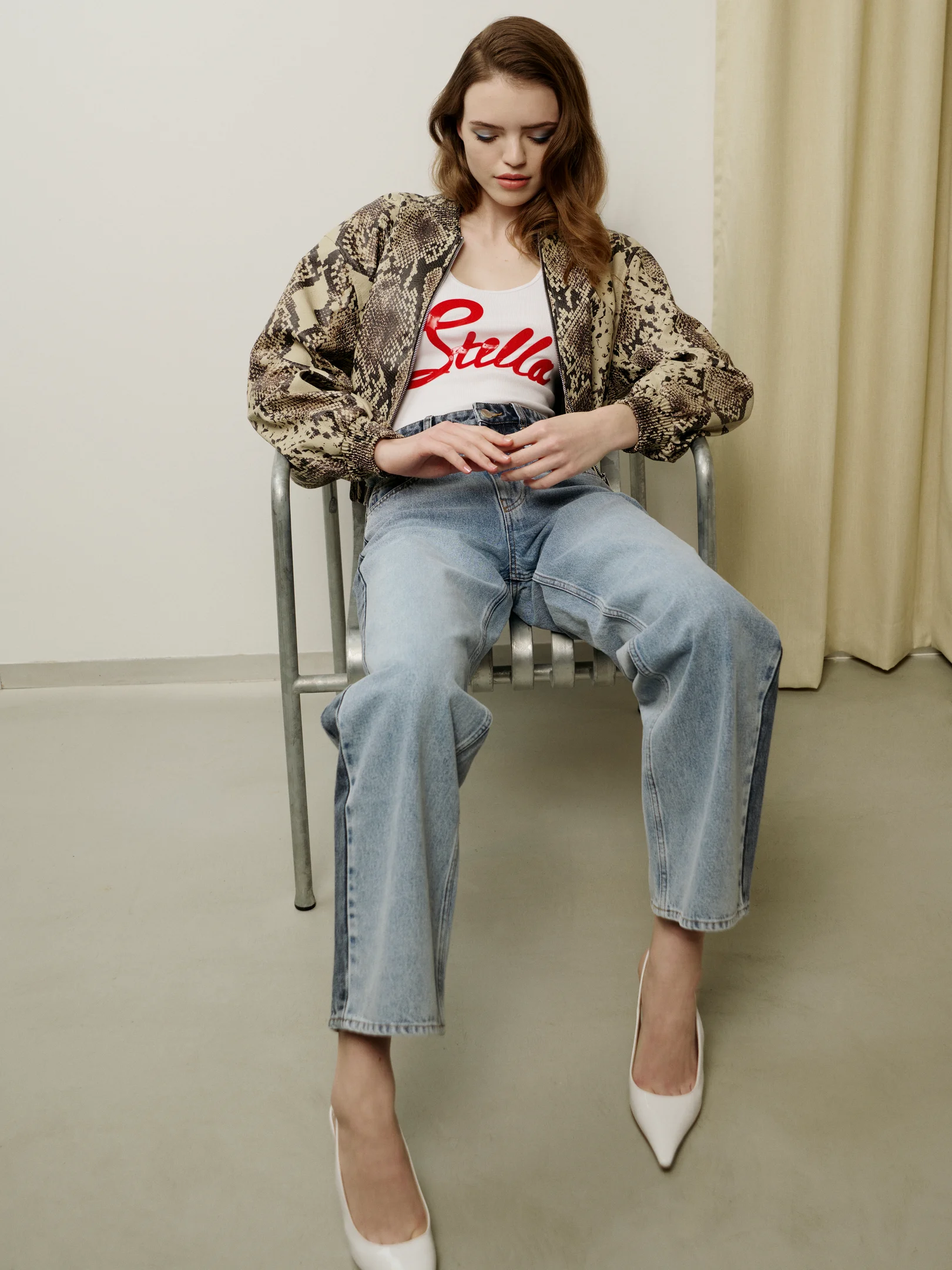 Frau sitzt auf Stuhl und trägt eine Bomberjacke mit Schlangenprint, weißes Stella-Top und weite Jeans in hellem Studio