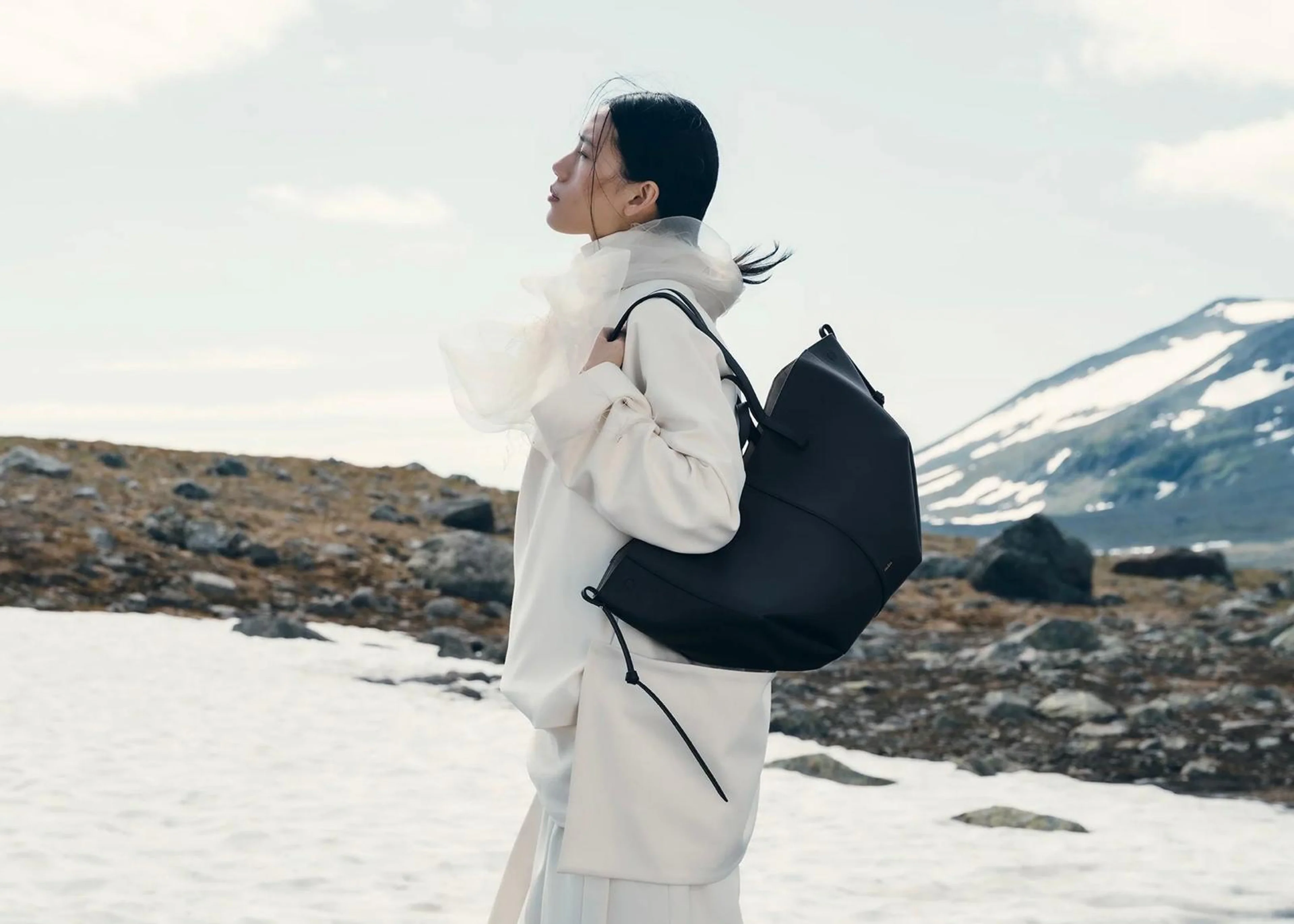 Frau in weißer Kleidung vor einer Bergkulisse mit Schnee, die eine schwarze, skulpturale Polène Handtasche auf dem Rücken trägt.